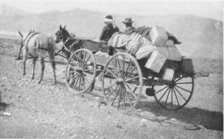 A black and white photo of two horses pulling a wagon filled with boxes and two passengers.