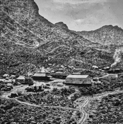 Black and white image of a cluster of buildings and canvas tents in a wide desert canyon.