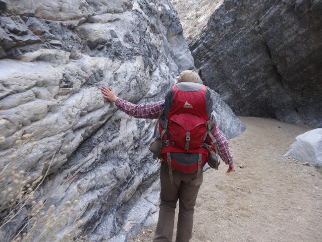 A backpacker touches the smooth rock wall of a vertical canyon as they walk away from the camera deeper into the canyon.