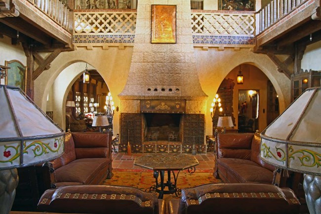 Lamps sit on the left and right of a wooden table in the foreground. Leather couches and a fireplace are in the background.