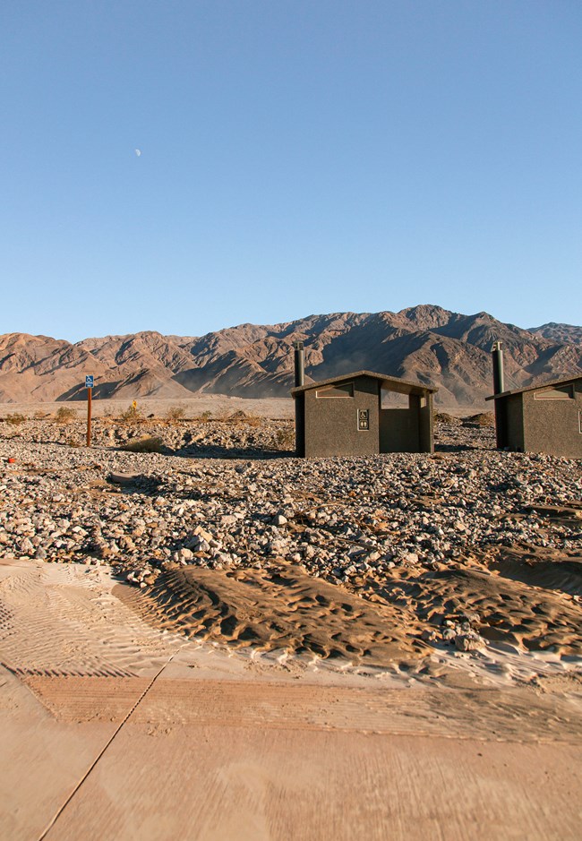 Two restroom structures sit in front of brown mountains, below large rocks carried from flash floods cover the parking lot.