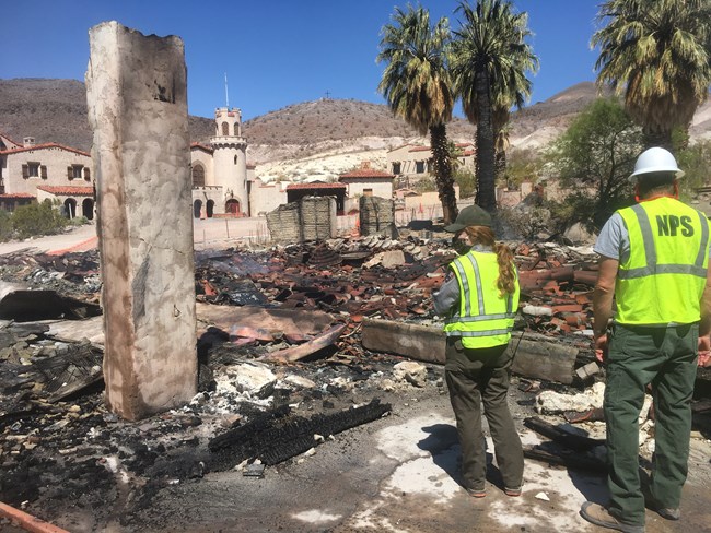 Two people wearing uniforms and reflective vest stand looking at a fully burned to the ground building.