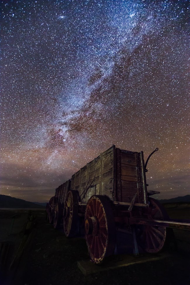 At night, a wooden wagon with large spoke wheels is seen from the front with the milky way and stars above.