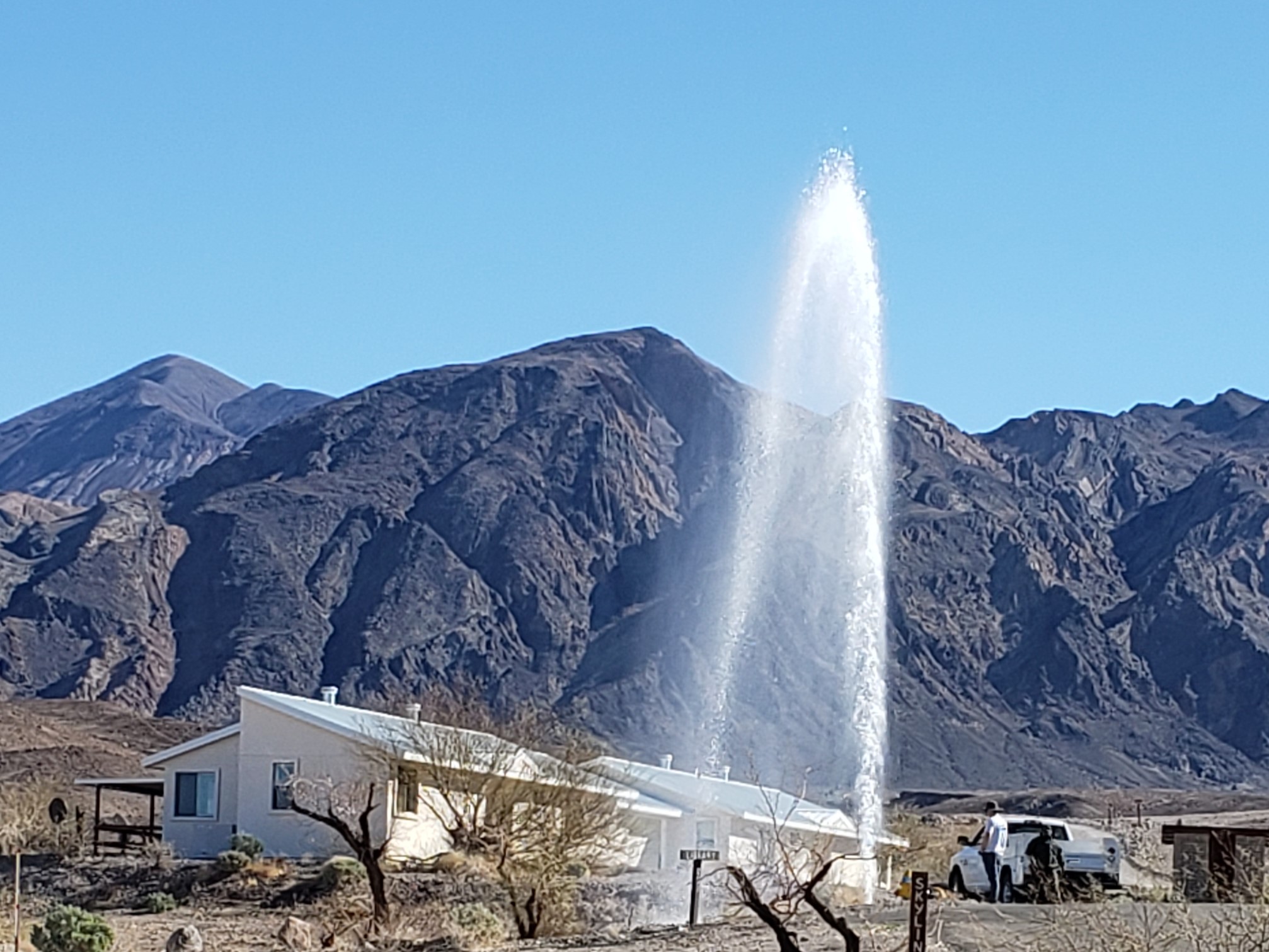 A white plume of water towers over the tan-color house behind it. Bare gray mountains and blue sky are in the background.