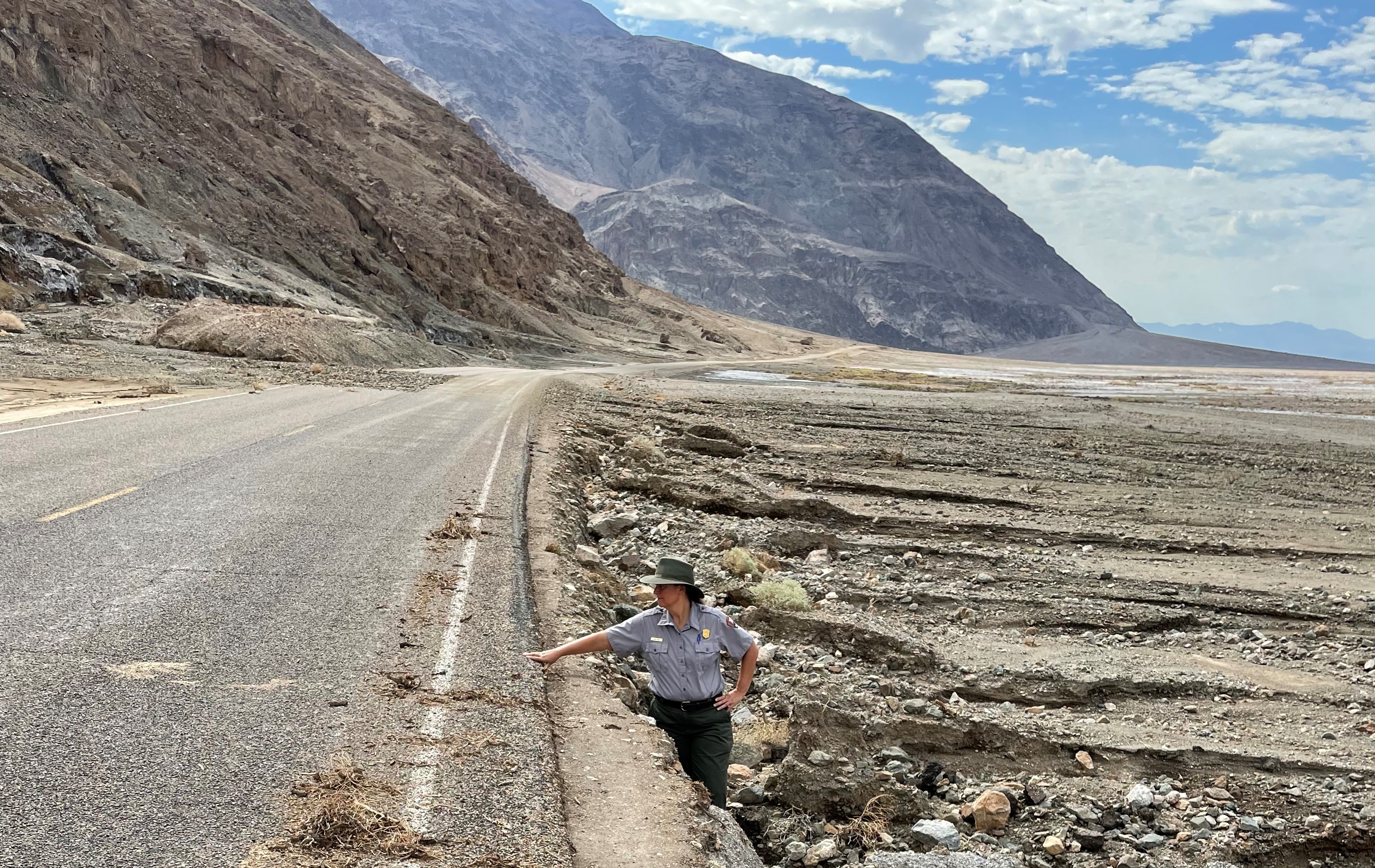 A woman in National Park Service uniform stands in an eroded road shoulder. Her arm is almost horizontal, touching the edge of the pavement.