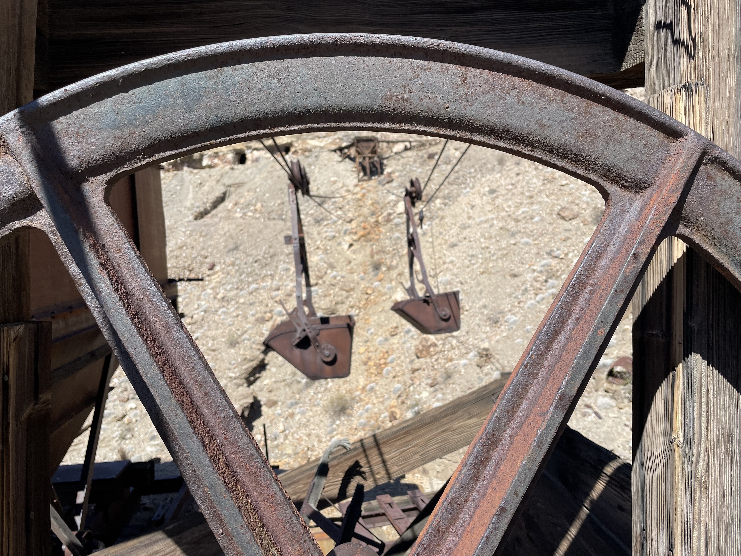 Spokes of metal equipment frame a view of metal ore buckets hanging from a cable in a desert landscape.
