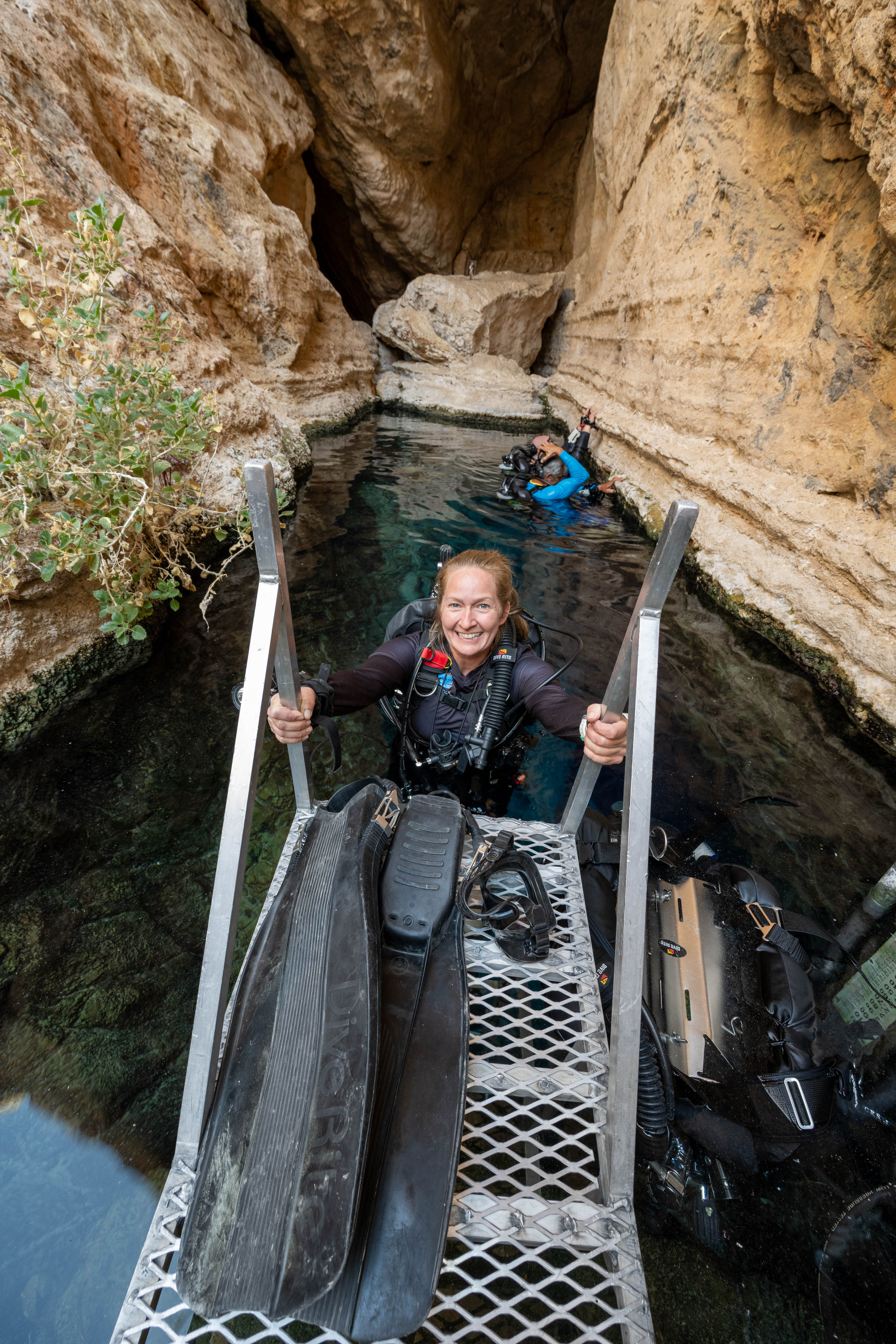 A woman smiles as she backs down into a narrow body of water from a metal platform. A diver in blue is in the background.