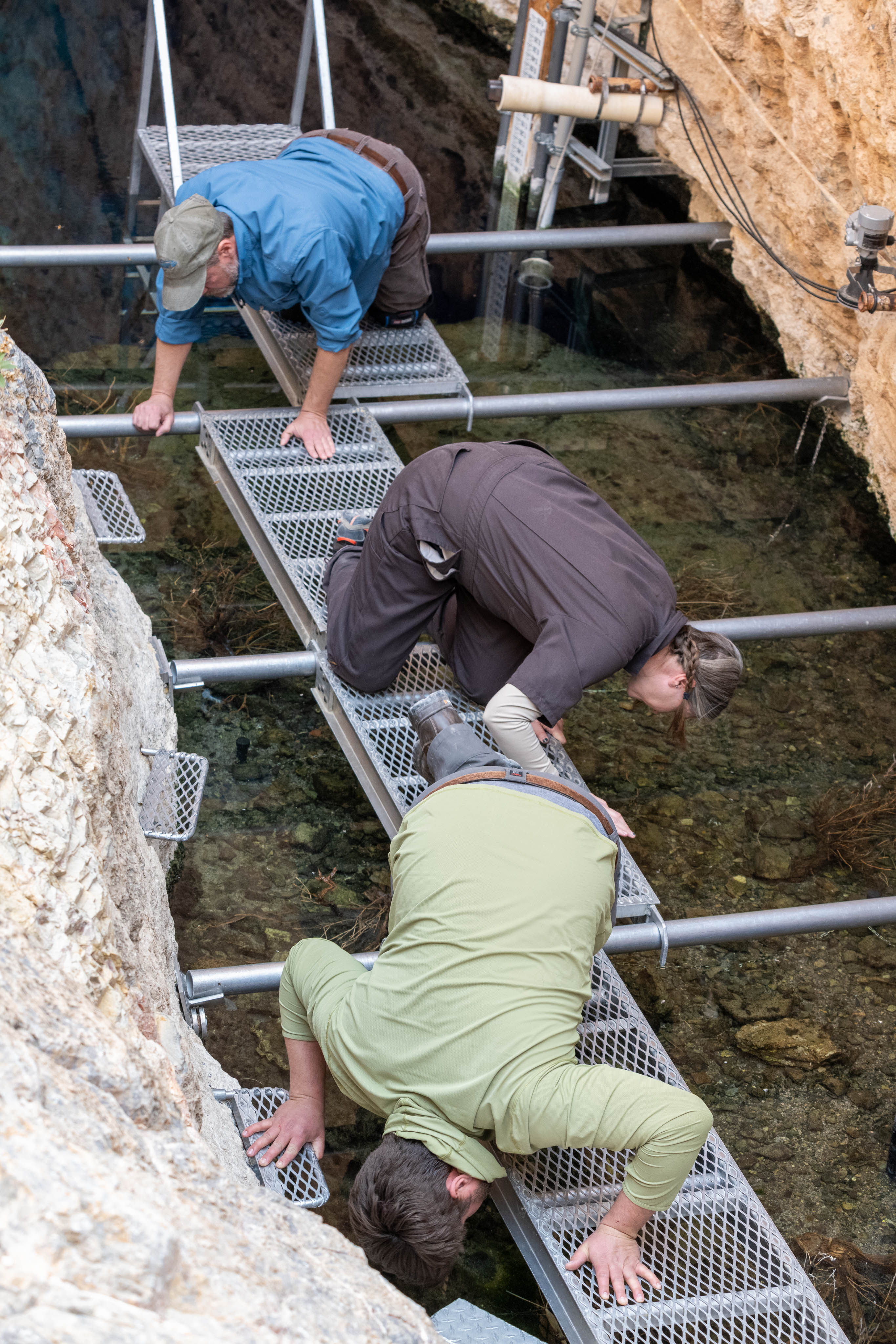 The image captures three people kneeling on metal grating platforms over water within a rocky crevice. Two men and one woman are closely examining the water below them.