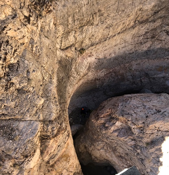This photograph is looking down from a helicopter. In the helicopter's landing bar and part of a person's shoe are in the lower right corner. The rest of the image is looking straight down into a narrow canyon. One bright orange spot is seen in the canyon