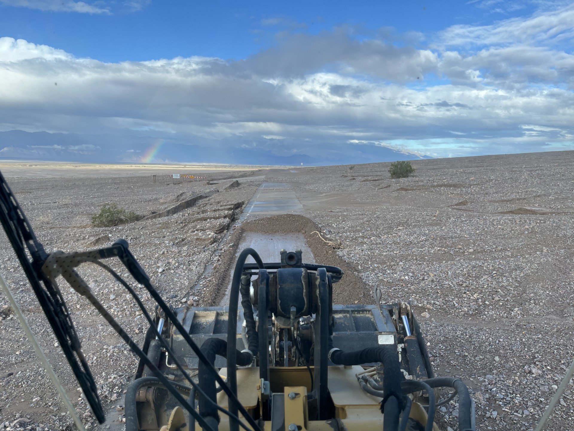 Looking out windshield at the front of yellow heavy equipment and a paved road partially covered with dirt. A rainbow is in the distance.
