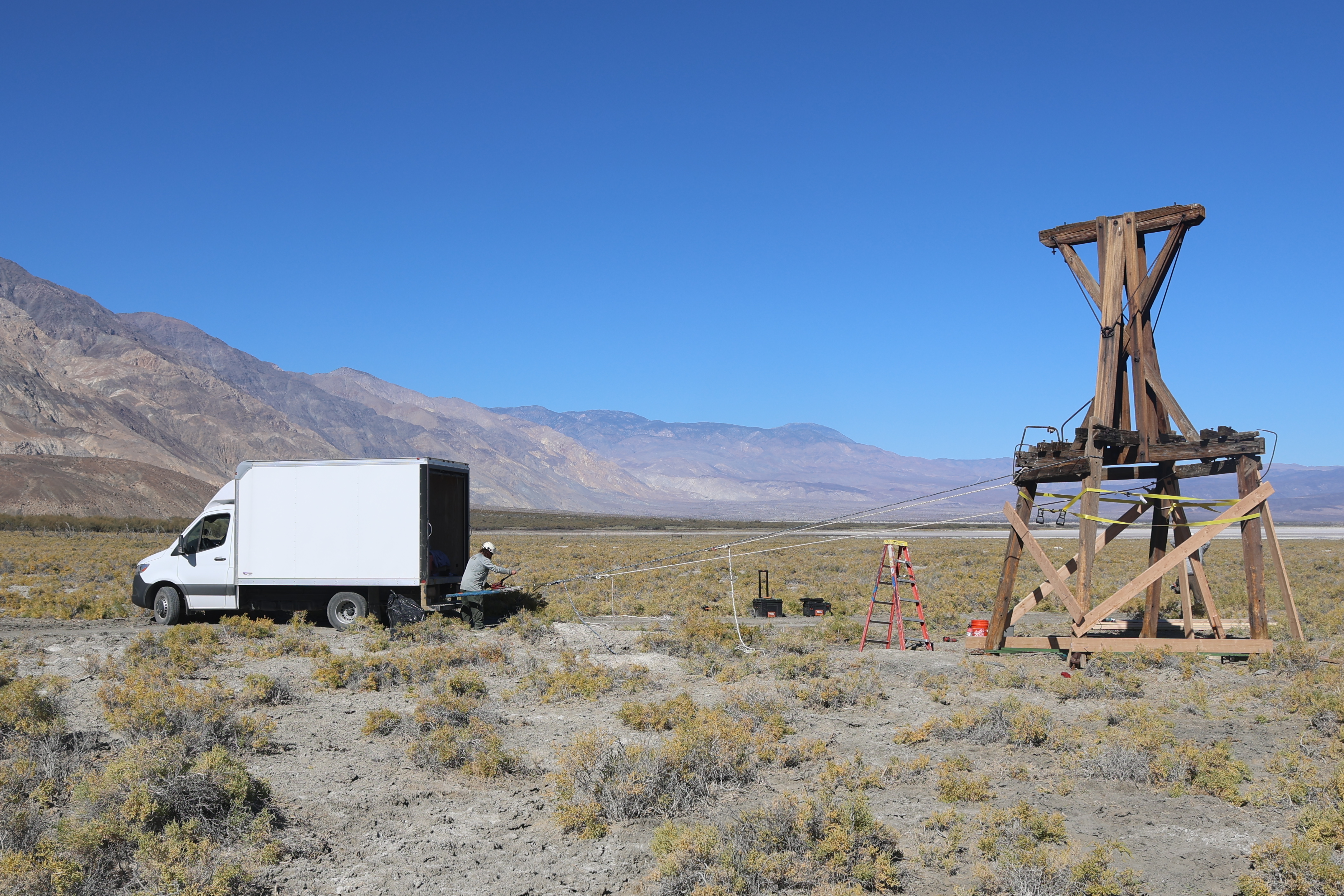 A white box truck is on the left.  An upright 20-foot wooden tower is on the right. A person operates a ratcheting device on white ropes that connect the truck to the tower.