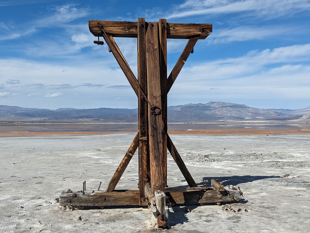 Wooden structure stands upright on flat white ground. A shallow lake and desert mountains are in the background.