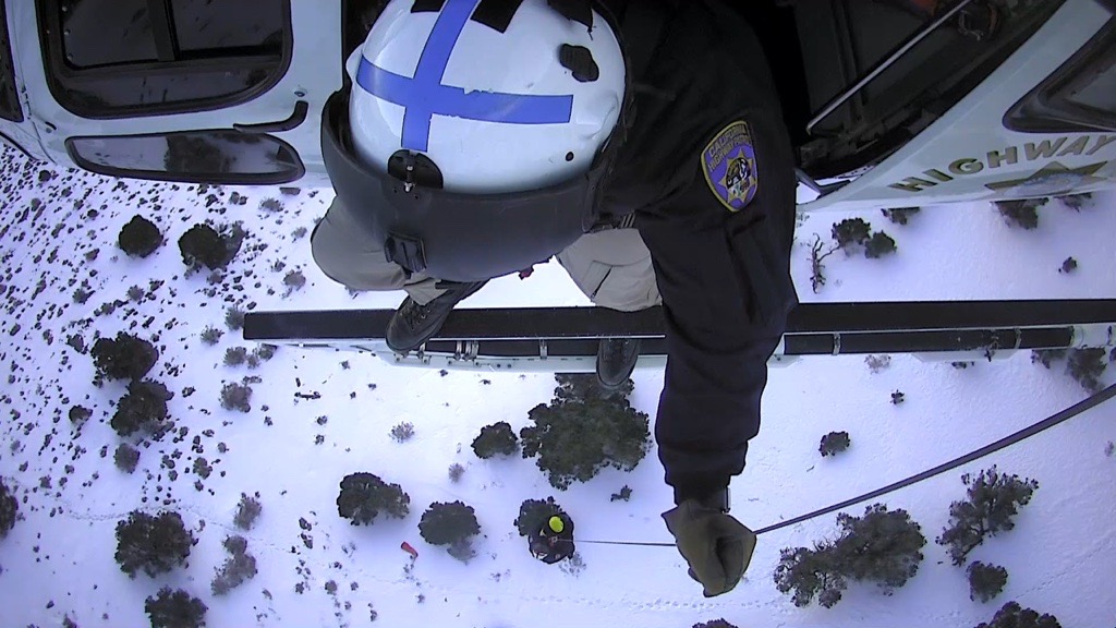 Image is taken looking straight down. The top of a person's helmet is seen sticking out the open helicopter door. A person hangs from a cable above a snowy landscape.
