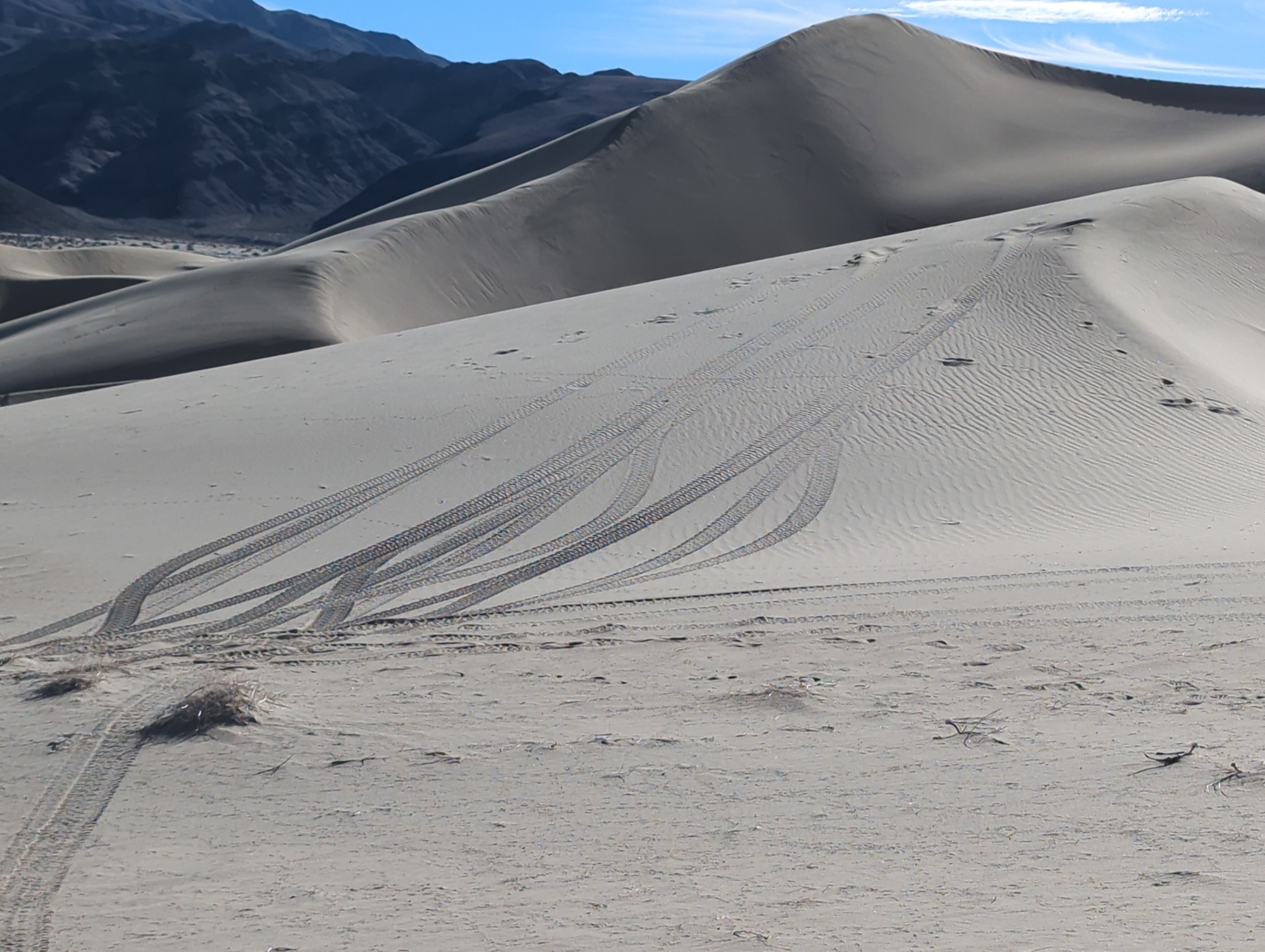 Vehicle tracks lead up a white sand dune from the left side of the image to the right side. Blue sky and dark mountains in the background.