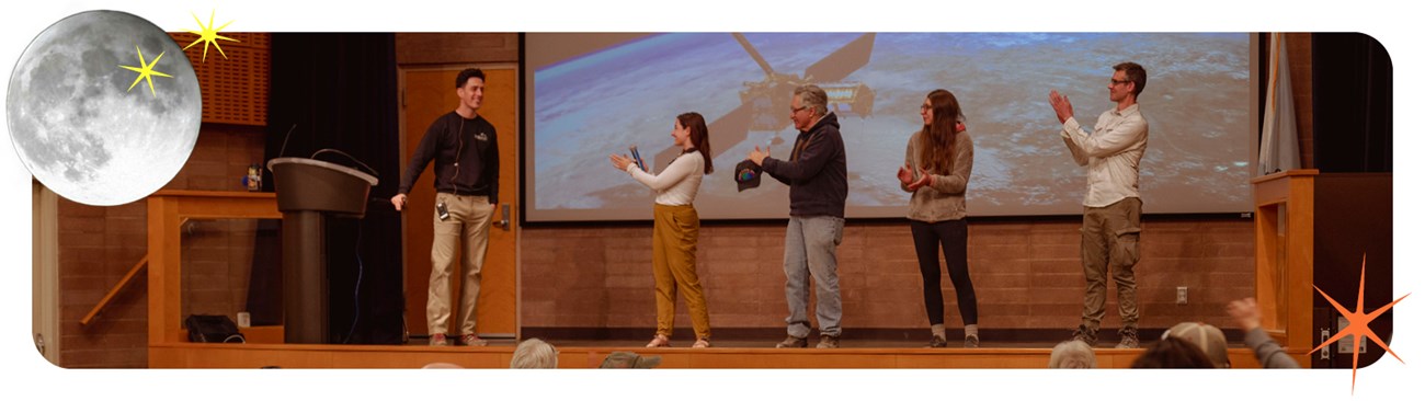 Five people stand on a wooden stage indoors in front of a screen.