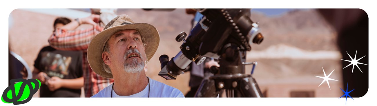 Man with gray beard and hat looks to the sky sitting next to a telescope.