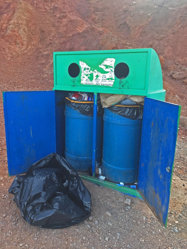 Two trash cans sit in an open box, with a full bag in front, in a desert landscape.