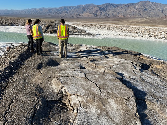 Three people standing on a raised dirt road looking at a washed out section covered with green water in a salty desert landscape.