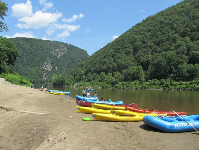 Kayaks and canoes line the shore of a sandy boat launch on the river. The Delaware Water Gap looms in the background covered in green foliage. 
