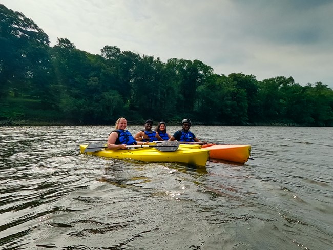 Four kayakers (two men, two women) face the camera with a row of thick trees lining the river bank behind.