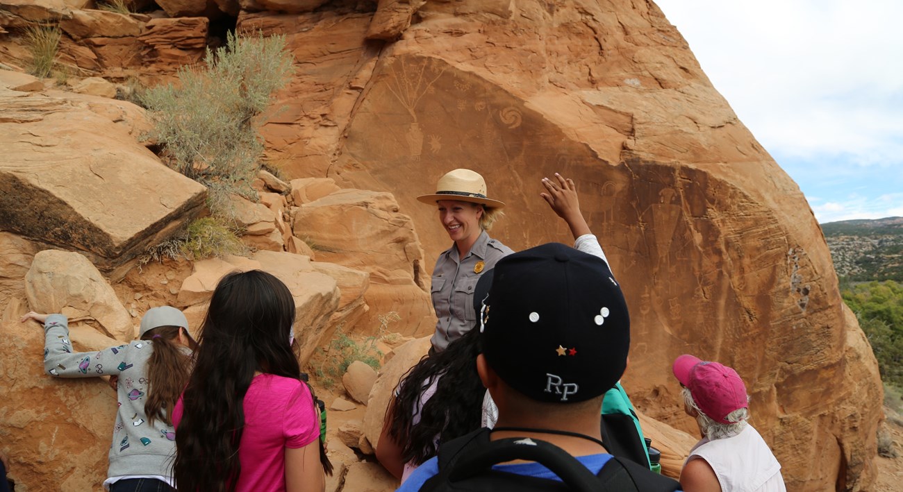 A smiling park ranger stands among students before a wall of petroglyphs.