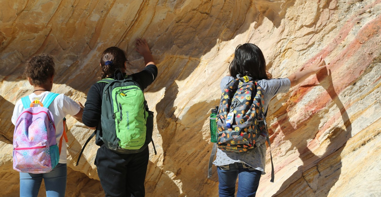 Students with backpacks on touch the side of a sedimentary rock face.