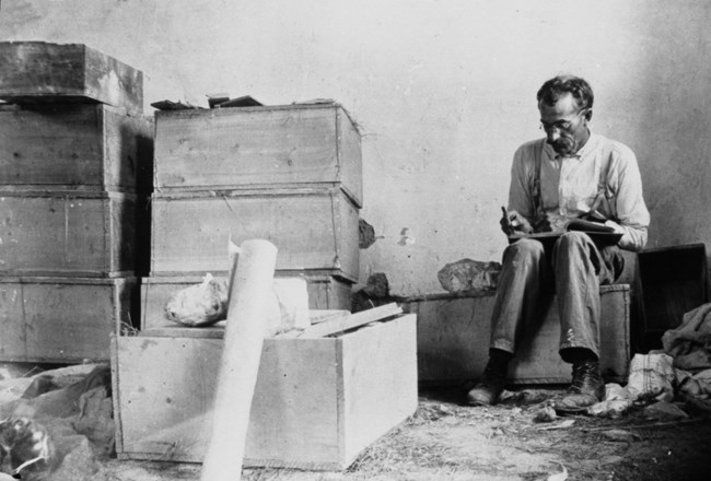 A black and white photograph of Earl Douglass surrounded by stacked crates. He is sitting down, with a notebook open on his lap, writing in it.