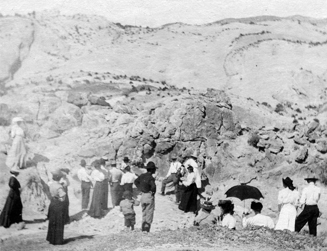 A black and white photo showing visitors of all ages at the Carnegie Quarry dig site. Children and women in dresses with umbrellas for shade look down at the original 8 Apatosaurus bones in the rock.
