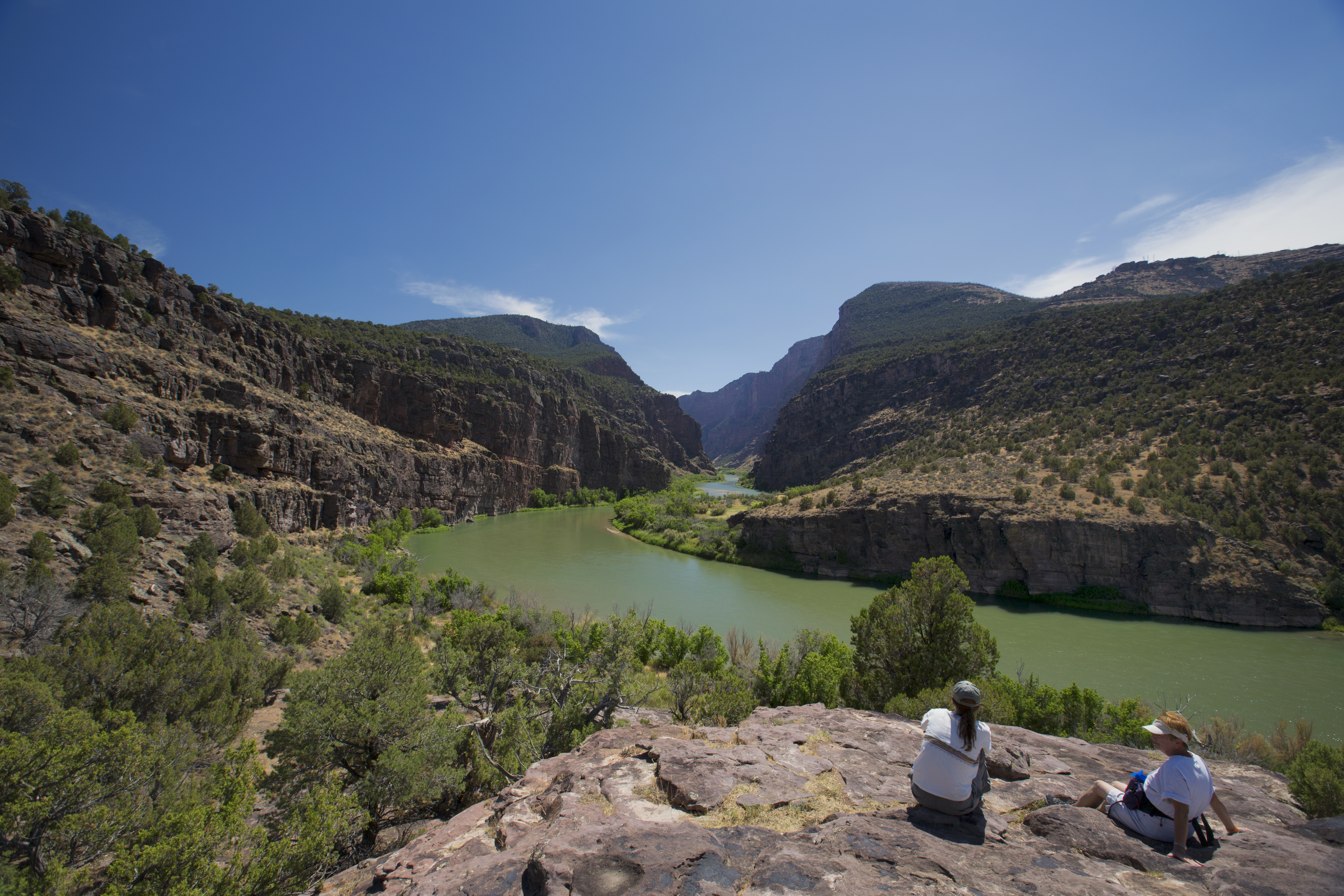 Two visitors sitting on a rock looking at the view of a river flowing into a canyon.