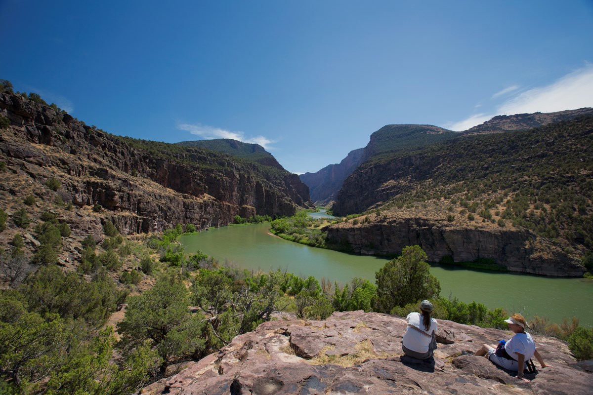 Two visitors sitting on a rock looking at the view of a river flowing into a canyon.