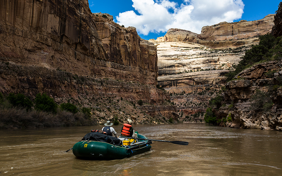 2 people in a green rubber raft float on a calm river as cliffs of yellowish tan rock tower above them