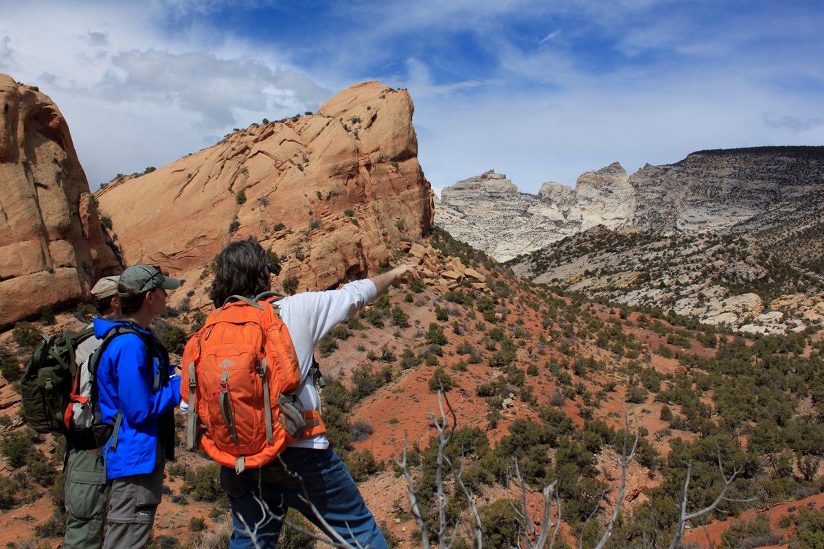 Three people, one of them pointing, stand in front of large orange-colored rocks.