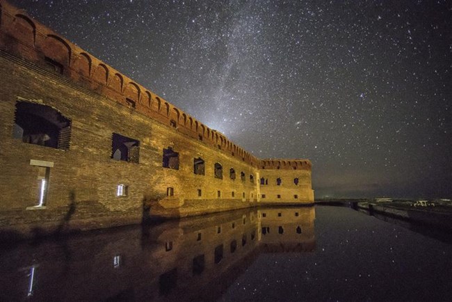 A large, brick fort and the stars of the Milky Way are reflected in the still waters of a moat.