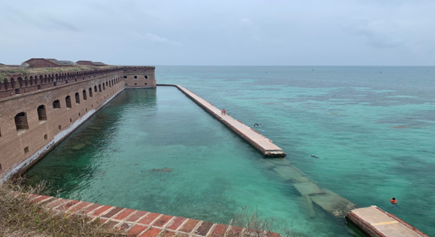 A view of the broken section of the moat wall in front of the brick fort with a view out to shallow green water