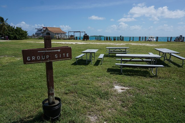 A wooden sign with "GROUP SITE" written on it on grass in front of picnic tables