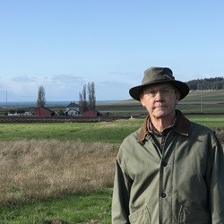 Man in hat in front of old farmstead.