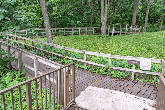 A wooden ramp goes up and into the woods. A sign on it reads "3 Mounds", indicating the conical mounds adjacent to the visitor center.