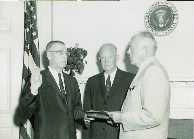 General Bragdon, standing left, takes an Oath of Office with his hand on a bible. President Eisenhower stands in center of photograph