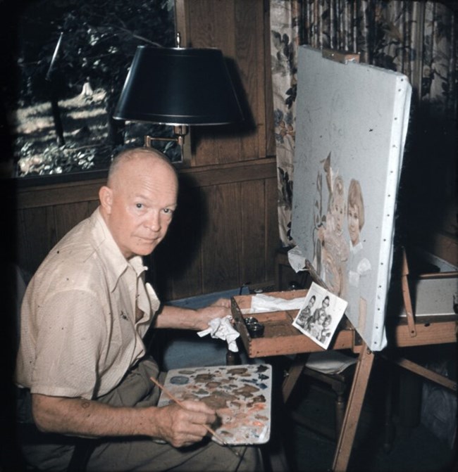A man sits at an easel with an array of paints and colors while working on a painting of two children.
