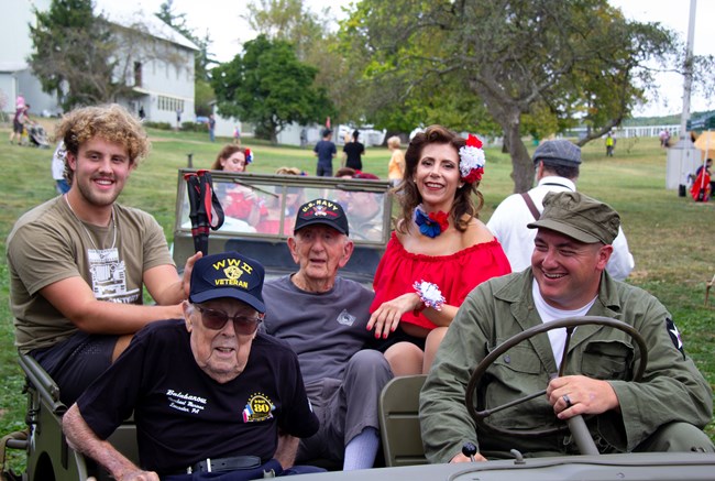 An image of several WWII veterans sitting in a green army jeep with WWII living history volunteers