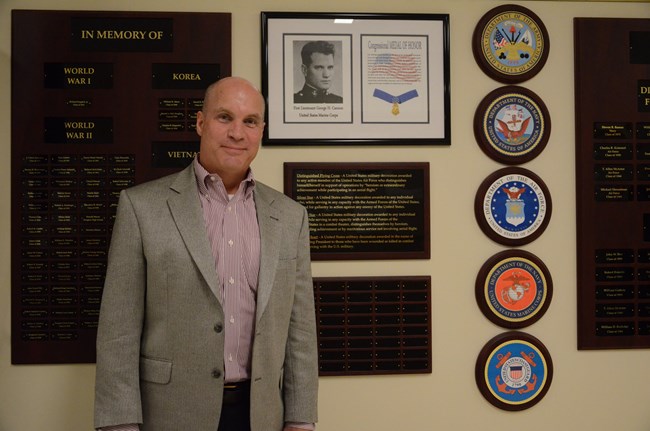 A man in a grey suit coat stands in front of a wall