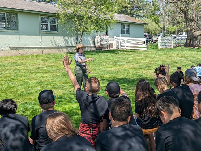 A park ranger speaks to a group of students underneath a tree on a sunny day