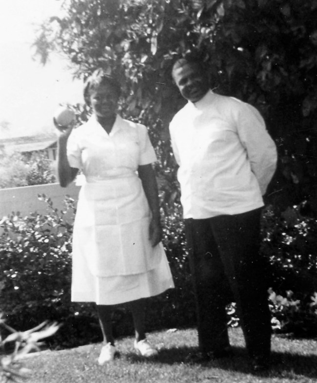 Delores and John Moaney smile for a photograph. Delores is wearing a white dress and holding up a ball in her right hand. John is wearing a white shirt and black pants; his arms are behind his back.