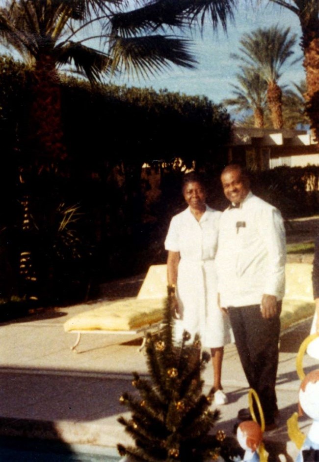Delores and John Moaney, both wearing white, stand and smile for a photograph. In the background are some palm trees and a large building.