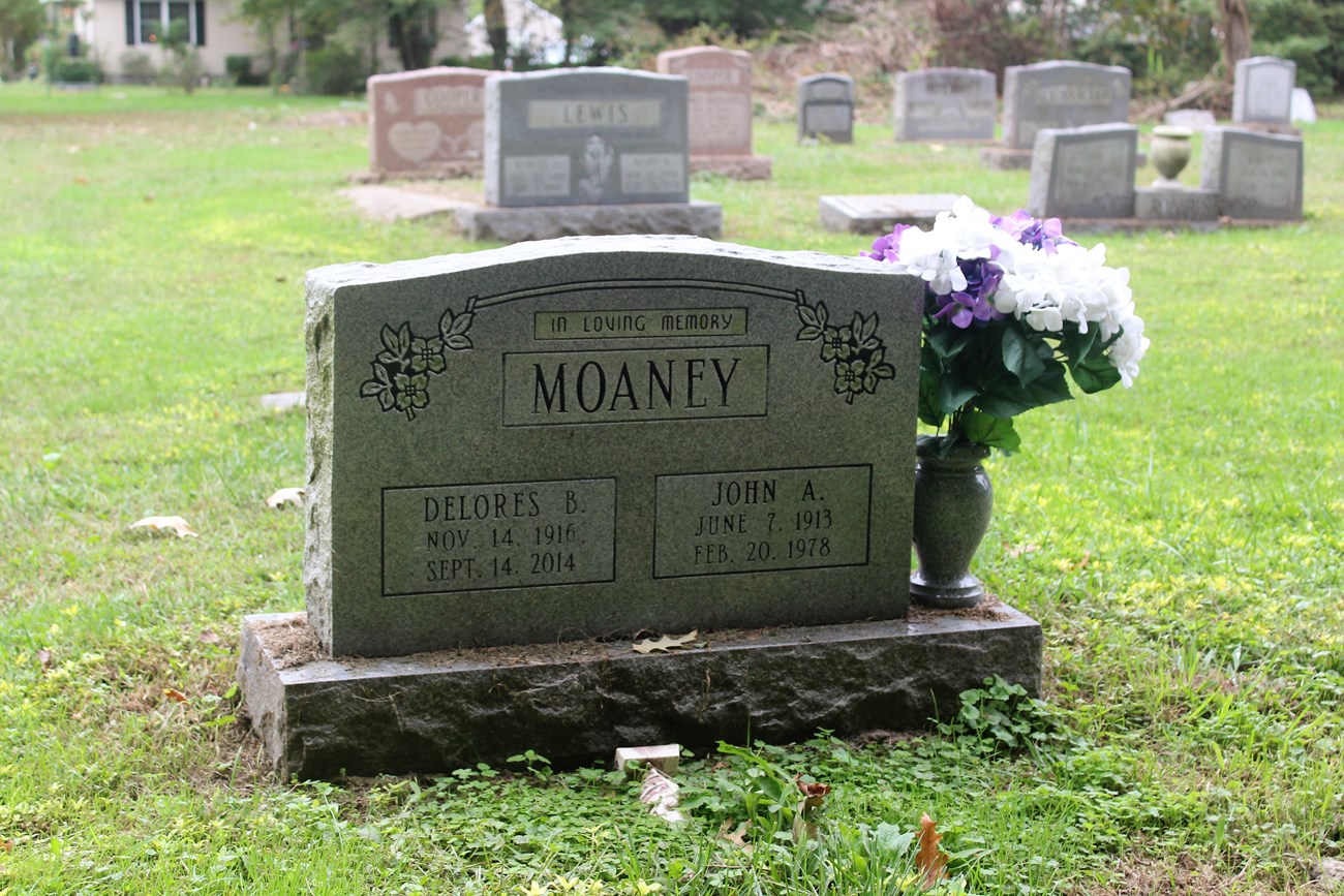 A granite headstone marks the grave site of John and Delores Moaney. An urn next to the grave has purple and white flowers.