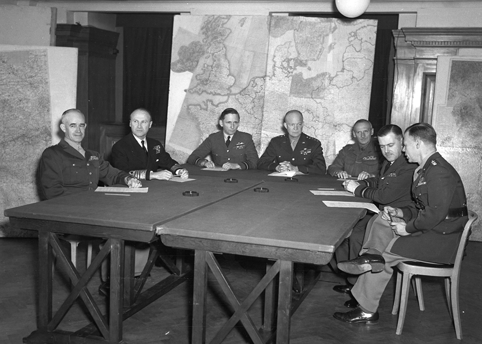 A black and white photo of seven American military officers in full uniform sit around a table discussing military operations. There are multiple large maps in the background.
