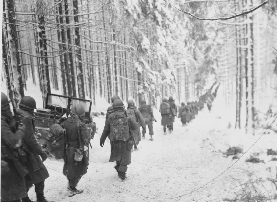 A black and white photo of American soldiers walking down a snow covered road in a forest during the Battle of the Bulge.