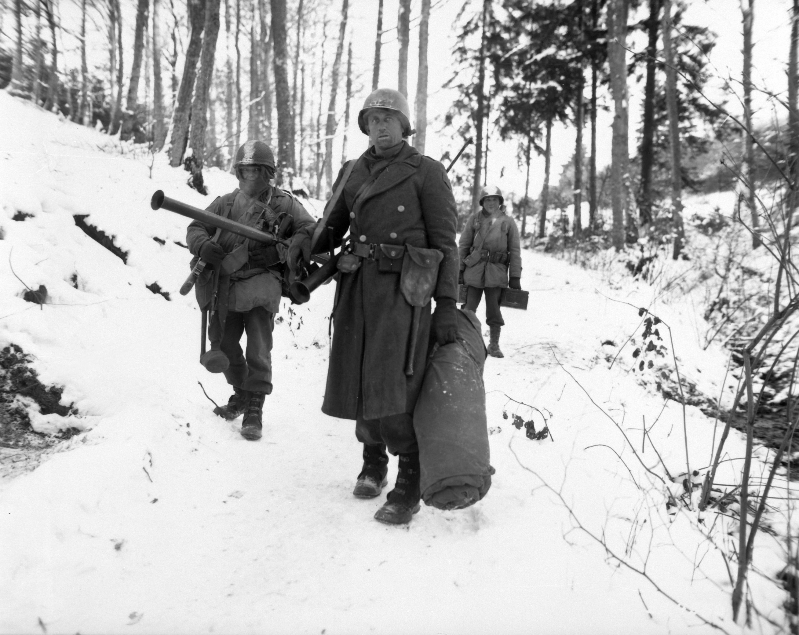 American World War II soldiers emerge from the woods. They are dressed in heavy coats and carry large sacks. There is snow on the ground.