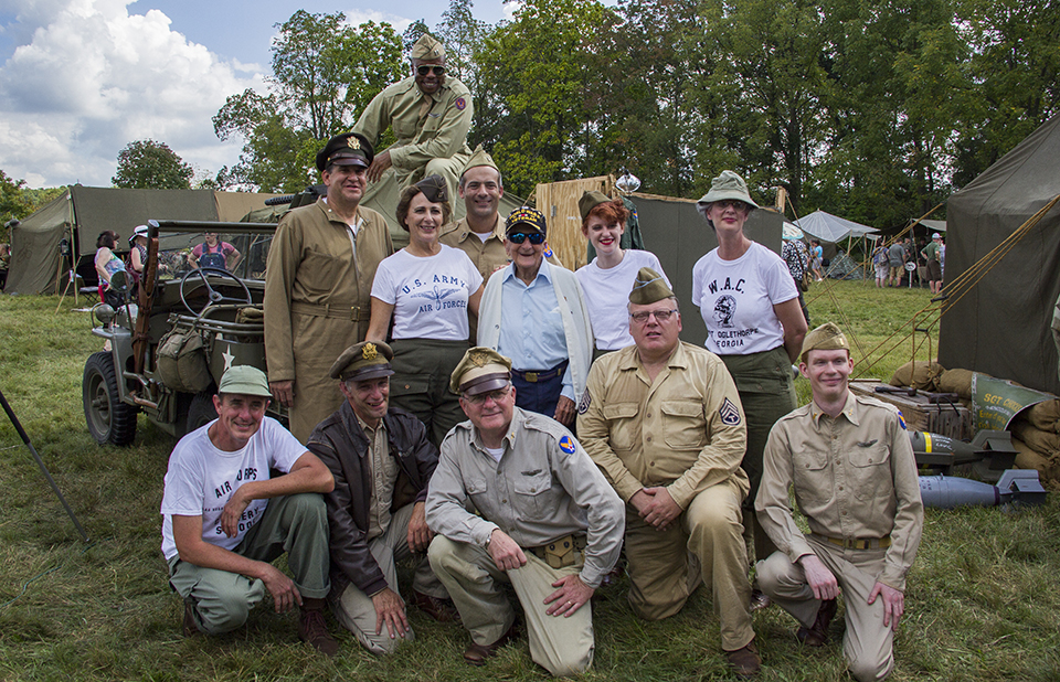 A group of World War II living historians pose for a picture with a World War II veteran. They are gathered around Jeep and camp tents are all around the group.