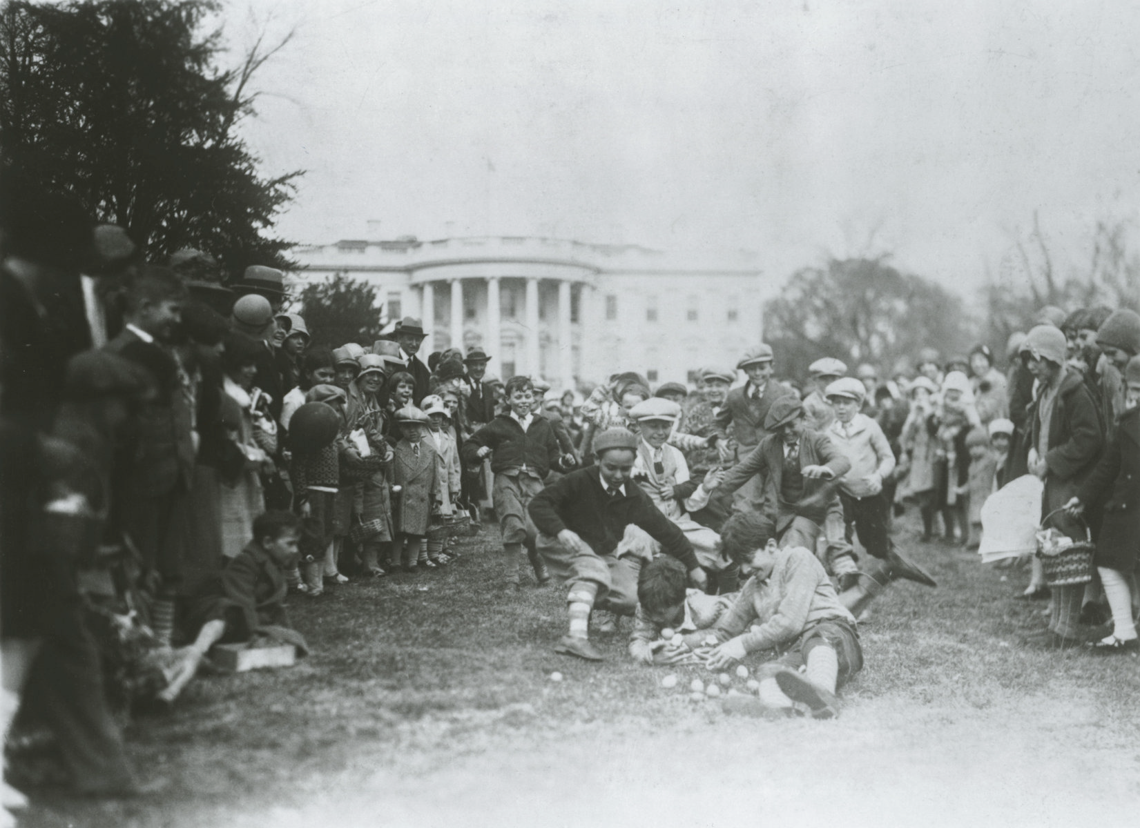 An old black and white photo of a group of young children running towards the center of the photo to gather Easter Eggs. The White House is in the background and a large group of onlookers surrounds the children.